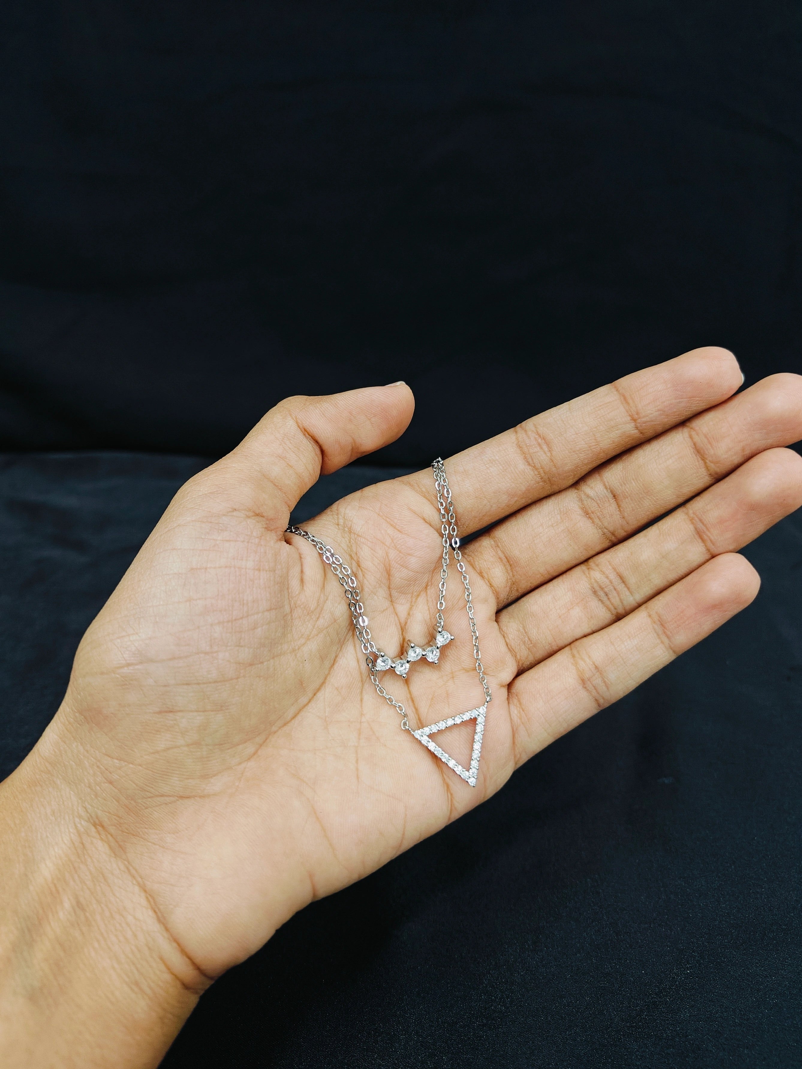 Hand holding a silver necklace with a triangle pendant against a black background